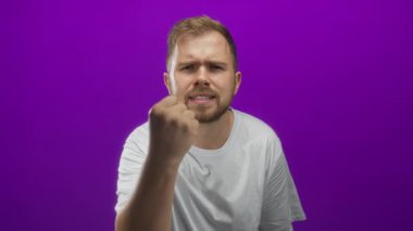 Yumruklarını sıkmış ve stüdyoda canlı mor bir arka plana karşı ellerini kaldırmış beyaz tişört ve sakal giymiş bir adam..