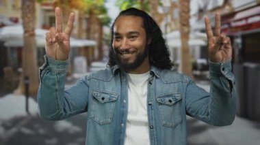 Gülümseyen adam, açık hava kafenin yanındaki sokakta mavi kot ceket ve beyaz tişört giyen barış işareti tutuyor; neşe barışı..