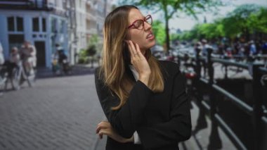 Meşgul bir şehir caddesinde kulak ağrısı için kulağına dokunan bir kadın. Kanal kenarında, çene çene ve somurtma;.