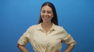 Mavi stüdyoda elleri kalçalarında gülümseyen, bej gömlek giyen, kendine güvenen bir kadın..