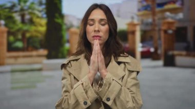 Şehir caddesinde gözleri kapalı dua eden bir kadın, bulanık şehir dekoru ve yeşillik ile çevrili, somut sükunet ve odaklanma..