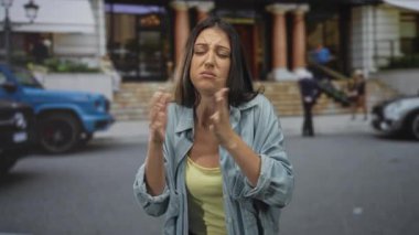 Elleri kelepçeli bir kadın bir binanın önünde bir sokak önünde yalvarıyor, gözleri kapalı ve kaşları çatık; umutsuzluk.