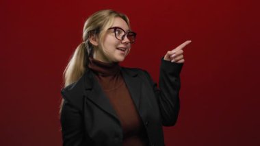 Gözlüklü ve ceketli bir kadın kırmızı bir duvara karşı ifadeyle işaret ediyor, profesyonel ve kendine güvenen bir tavır sergiliyor..