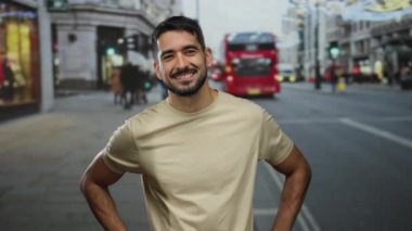 Kentin arka planında, sembolik kırmızı otobüslerle güvenli bir şekilde duran genç adam büyüleyici bir gülümseme ve kozmopolit bir şehir sahnesinde rahat bir duruş sergiliyor.