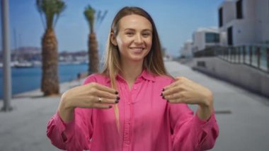 Pembe tişörtlü kadın, arka planda palmiye ağaçları ve suyla güneşli bir sahil yürüyüşünde gülümsüyor, deniz kenarında bir yerde neşe ve rahatlama yaşıyor..