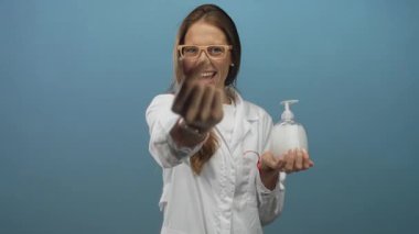 Beyaz laboratuvar önlüklü kadın bilim adamı sabun makinesini tutuyor ve mavi stüdyoda eli ile parmak kalbi gösteriyor; hijyen tanıtım özgüveni.