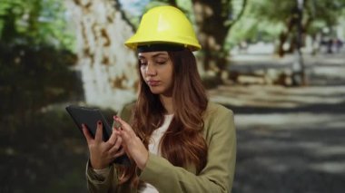 Güneşli bir günde parkta tablet kullanarak açık hava ortamlarında modern teknolojiyi gösteren bir şapka takan genç İspanyol kadın mühendis..