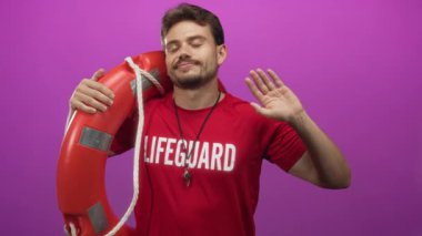 Kırmızı bir yüzük ve ıslık tutan genç bir cankurtaran, gülümseyen ve bir elini mor stüdyoda sallayan; gurur görevi.