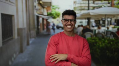 Kırmızı kazaklı yakışıklı bir adam şehir caddesindeki terasta gülümsüyor, sıcaklık ve cazibe saçıyor..