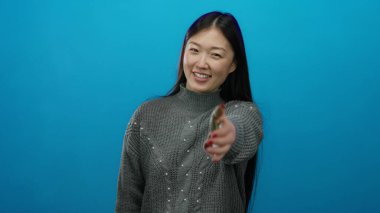 Mavi arka planda el sıkışmak için gülümseyen bir kadın, rahat bir kazak giyiyor, dostça bir hareket sergiliyor..