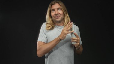 Man with long blond hair in gray shirt carefully inspects and opens white waterbottle cap near lips at studio; thirst.
