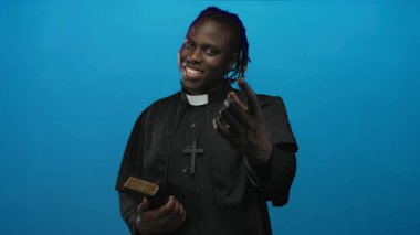Papaz tasması takan ve elinde İncil olan bir adam mavi stüdyodaki kamerayı işaret ediyor..