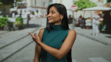 Güneşli caddede gülümseyen bir kadın yeşil elbiseli, açık hava kafeleri ve bulanık yürüyen insanlarla dolu şehir manzaralı bir yerde güven içinde yol gösteriyor..