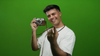 İspanyol asıllı genç bir adam elinde bir kamera tutuyor ve canlı yeşil bir duvar arkaplanına karşı bir jest yapıyor, büyüleyici ve davetkar bir etkileşim..