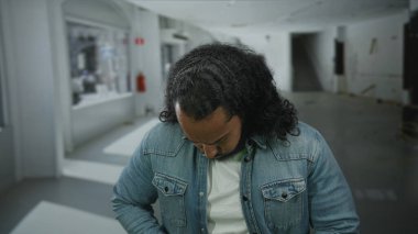 Sakallı ve uzun saçlı bir adam göğsünü sıkıyor ve boş stüdyo odasında gözlerini kapatıyor, kot ceket görünür, eli göğüs kemiğine bastırılmış, muhtemel kalp krizi; acı dayanıksızlığı iyileşme çabası.