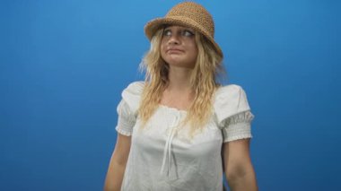 Mavi stüdyoda, saman şapka ve beyaz bluz giyen bir kadın işaret ediyor ve kollarını çapraz yapıyor..