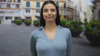 İspanyol kadın sokakta dikiliyor, açık havada gülümsüyor. Şehir arka planında rahat moda, kendine güvenen ifade, canlı bir yaz günü.