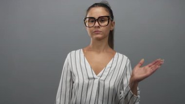Çizgili bluz giyen ve büyük gözlüklü bir kadın. Gri stüdyo ortamında elini kaldırıyor ve avucunu açıyor. Neşeli..