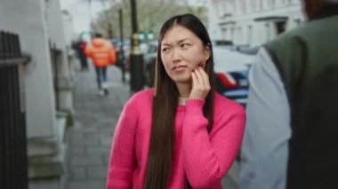 Woman touching her cheek with a pained expression in a city street, wearing a bright pink sweater, surrounded by urban activity and blurred pedestrians in the background.