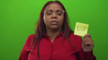 Kadının elinde kırmızı gömlek ve yuvarlak gözlük takan açık yeşil bir stüdyoda, üzerinde vergi metni bulunan sarı yapışkan bir not var..