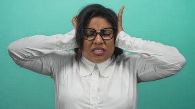 Gözlüklü kadın, yeşil arka planda yaşadığı hayal kırıklığını ifade eden kulaklarını kapatıyor. Bu da ofis ortamında stres olduğunu gösteriyor.