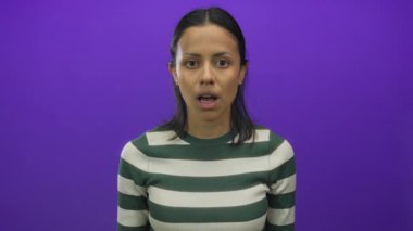 İspanyol kadın, yeşil çizgili gömleği olan, izole edilmiş mor bir arka plan duvarında dururken şaşkınlığını dile getiriyor..