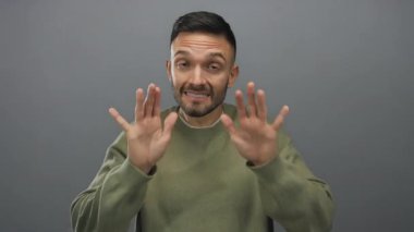 Elleri gri arka planda, sakin hareketler yapan İspanyol bir adam, sıradan yeşil bir kazak giyiyor, tarafsız bir ortamda sükunetini ve güvenini ifade ediyor..