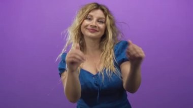 Sarı saçlı, mavi elbiseli bir kadın stüdyoda yüzü mora yakın bir kalp oluşturur..