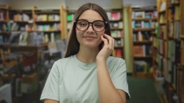 Genç bir kız kütüphane binasındaki ahşap kitaplıklar ve kitapların arasında gözlüklerine dokunur..