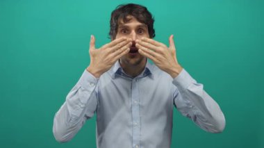 Açık mavi gömlekli genç adam, açık kollu jest yapıyor ve stüdyoda yeşil arka plana karşı ağzını açıyor..