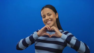 Hispanik kadın gülümseyen ve kalp şeklini oluşturan elleri ile canlı mavi izole arka plan, mutluluk ve pozitifliği ifade ediyor.