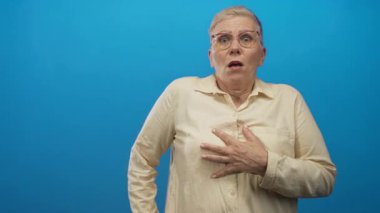 Göğüs göğüse, geniş gözlü ve ağzı açık, gözlüklü ve bej gömlekli bir kadın, canlı mavi stüdyo duvarına karşı duruyor; sürpriz tepki.