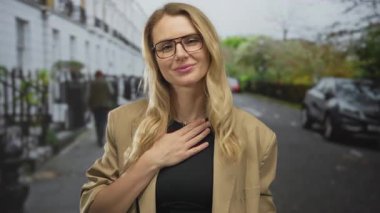 Sarı saçlı bir kadın, şehir caddesinde göğsünde sıradan giysilerle gülümsüyor, açık hava ortamında park etmiş arabalarla ve yeşilliklerle şükranlarını sunuyor..