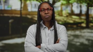 Güneşli bir günde dışarıda dikilen kırmızı gözlüklü ve örgülü saçlı bir kadın, şehir sokağında hoşnutsuzluğunu ifade ediyor kolları bağlı ve ciddi bir yüz ifadesi var..