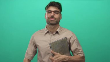 Gözlüklü gülümseyen adam kitabı tutuyor ve parlak yeşil stüdyoda el hareketleriyle kabul ediyor..