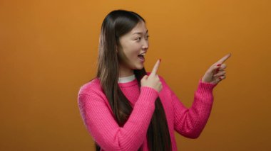 Parlak pembe bir süveter giyen, parlak turuncu arka planda gülümseyen ve işaret eden bir kadın canlı ve neşeli bir ifade sergiliyor..