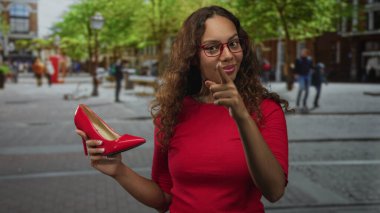 Kadın, kentin caddesinde kırmızı topuklu ayakkabıyı tutarken kamerayı işaret ediyor; kendine güven sağlamlığı.