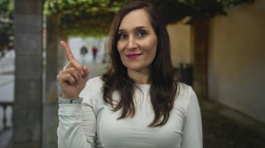 Yapraklı arka planı olan, kendine güvenen bir kadın, gençlik enerjisi ve İspanyol mirası sergiliyor, güneşli gökyüzünün altında gülümserken pozitifliğini ifade ediyor..