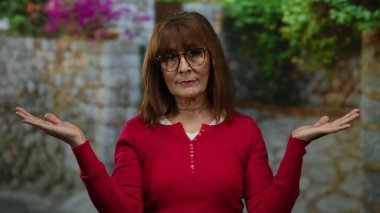 Kırmızı kazaklı, gözlüklü bir kadın sakince poz veriyor şehir sokağında bitkilerle, sükunet ve sükuneti temsil ediyor..