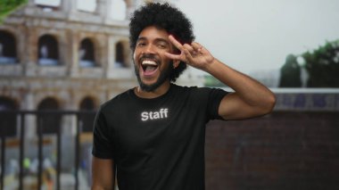 Roma 'nın ikonik kolezyumunda zafer işareti yapan gülen adam, canlı bir şehir arkaplanı sergiliyor..