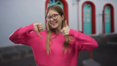 Pembe kazak giyen genç sarışın kadın, şehir caddesinde baş parmak kaldırma ve baş parmak kaldırma hareketi yapıyor; belirsizlik belirsizliği seçimi.