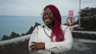 Bir kumsalda çapraz kollarıyla güvenle gülümseyen bir kadın bulutlu bir gökyüzünün altında deniz manzaralı, gözlüklü ve beyaz bir süveter giyen, açık hava manzaralı deniz manzaralı bir tatil atmosferi öneriyor..