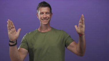 Eller havada, kameraya bakan, mor stüdyoda zeytin tişörtü giyen, gülümseyen, kendine güveni açık bir adam..