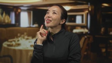 Elleri çenesinde gülümseyen ve restoran yemek odasında yukarı bakan, siyah kazak giyen ve bileğine akıllı seğiren bir kadın; düşünceli iyimserlik.