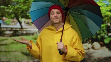 Bahçede gökkuşağı şemsiyeli sarı yağmurluklu genç adam arka planda ağaçlı palmiyeleri gösteriyor.