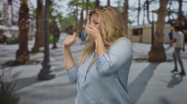 Palmiye ağaçlarıyla kaplı bir sokakta ağzı eliyle kaplayan ve başparmağını omzuna dayayan bir kadın şok reaksiyonu.