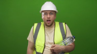 Stüdyoda elinde su şişesi tutan ve yeşil arka planlı yelek giyen mühendis sürpriz ve güvenlik endişesi.