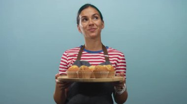 Kadın bir tepsi kek tutuyor ve onları görünür eller ile sunuyor bir deniz mavisi stüdyosunda; ev yapımı pişirme memnuniyeti.