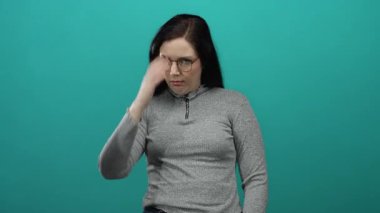 Gözlüklü bir kadın, yeşil bir arka plana karşı düşünceli bir şekilde el kol hareketi yaparak, elleri ve gözleri kapalı, dalgın bir yüz ifadesi sergiliyor..