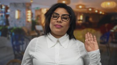 Restoran terasında gülümseyen gözlüklü ve beyaz gömlekli bir kadın şehrin arka planında neşeli bir güven gösterisi yapıyor..